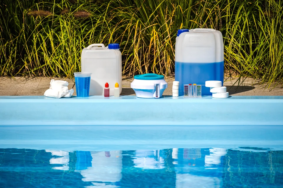 Gros plan sur une main tenant un kit d'analyse de l'eau sur une piscine claire et bleue, avec un équipement de nettoyage de piscine visible en arrière-plan